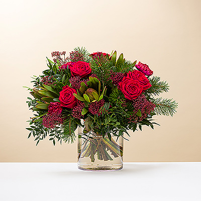 Deséales una Feliz Navidad con un precioso ramo rojo y verde. Las clásicas rosas rojas y otras flores se complementan con ramitas de pino y vegetación en este festivo arreglo floral navideño.