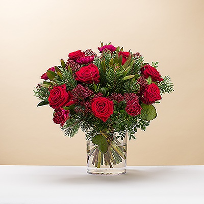 Deséales una Feliz Navidad con un precioso ramo rojo y verde. Las clásicas rosas rojas y otras flores se complementan con ramitas de pino y vegetación en este festivo arreglo floral navideño.