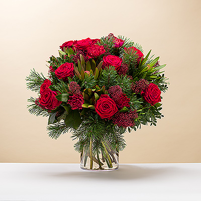 Wünschen Sie ihnen ein frohes Weihnachtsfest mit einem wunderschönen roten und grünen Strauß. Klassische rote Rosen und andere Blüten werden durch Tannenzweige und Grün ergänzt - ein festliches Weihnachtsgesteck.